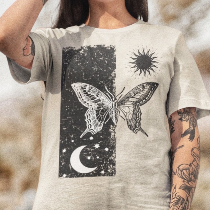 May include: A light gray t-shirt with a black and white graphic print of a butterfly, a crescent moon, and a sun. The graphic is on a distressed background.