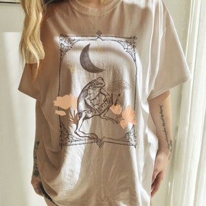 K&ouml;nnte beinhalten: Ein hellbraunes Oversize-T-Shirt mit einem grafischen Aufdruck einer unter einem Halbmond sitzenden Kr&ouml;te. Die Grafik ist mit einem dekorativen Rahmen versehen und enth&auml;lt Blumen und Sterne.