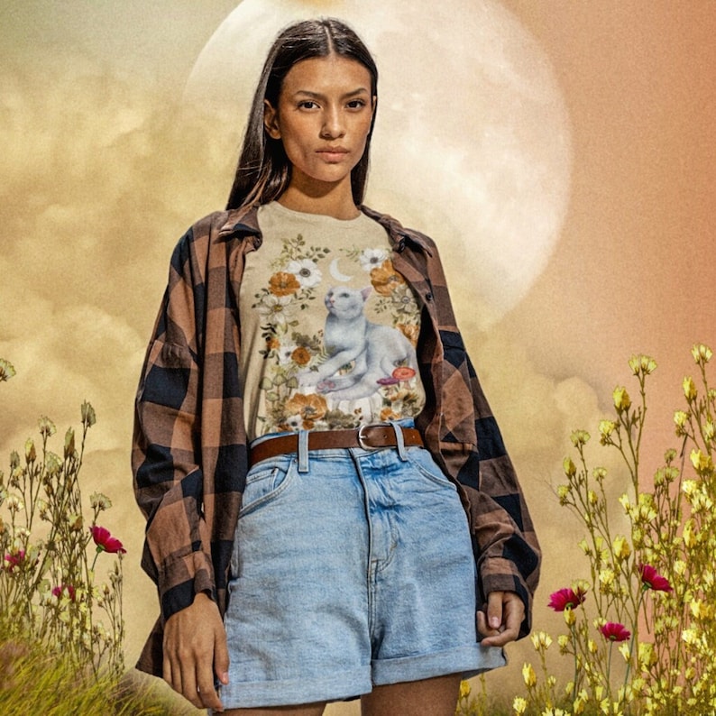 May include: A woman wearing a brown and black checked shirt over a white t-shirt with a floral print and a white cat illustration. She is also wearing blue denim shorts and a brown belt.
