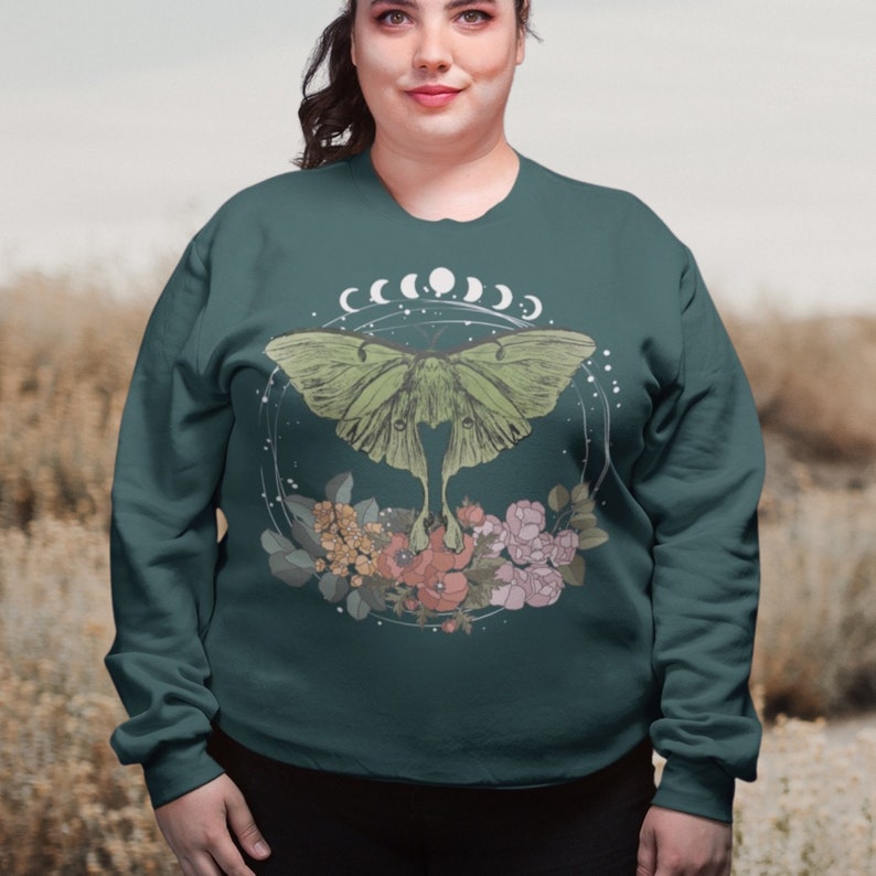 May include: Teal blue sweatshirt with a green luna moth graphic, surrounded by white crescent moons and pink and yellow flowers.