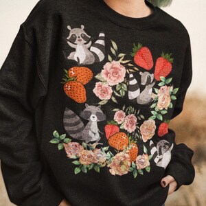 Pu&ograve; includere: Felpa nera con una stampa grafica di procioni e fragole circondate da fiori. I procioni indossano una corona di fiori e tengono delle fragole.