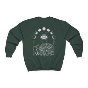 Op de afbeelding: Donkergroene sweatshirt met een witte afbeelding op de rug. De afbeelding bevat een oog, maanfasen en bloemmotieven. Tekst aan de linkerkant luidt "what you think you become" en aan de rechterkant "what you feel you attract."
