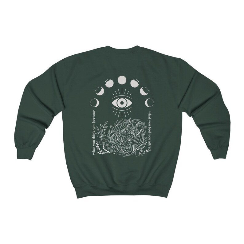 Op de afbeelding: Donkergroene sweatshirt met een witte afbeelding op de rug. De afbeelding bevat een oog, maanfasen en bloemmotieven. Tekst aan de linkerkant luidt "what you think you become" en aan de rechterkant "what you feel you attract."