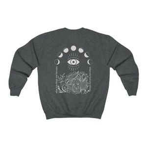 Op de afbeelding: Donkergrijze sweatshirt met een witte afbeelding op de rug. De afbeelding bevat een oog, maanfasen en botanische elementen. Tekst aan de linkerkant luidt "wat je denkt word je" en aan de rechterkant "wat je voelt trek je aan."