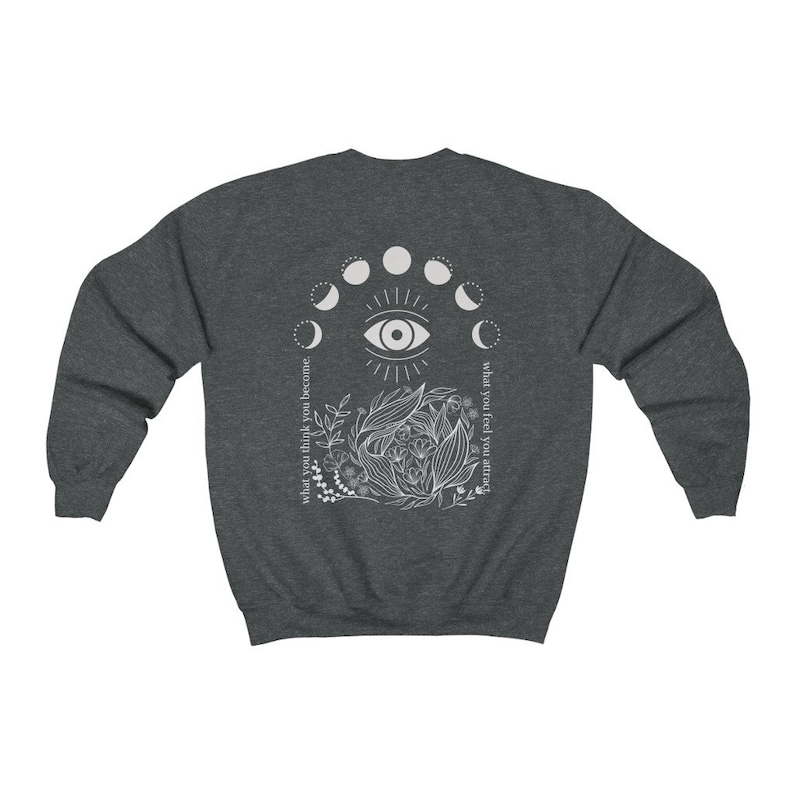 Op de afbeelding: Donkergrijze sweatshirt met een witte afbeelding op de rug. De afbeelding bevat een oog, maanfasen en botanische elementen. Tekst aan de linkerkant luidt "wat je denkt word je" en aan de rechterkant "wat je voelt trek je aan."