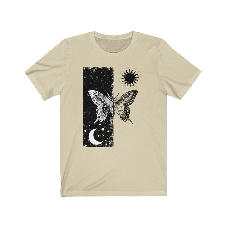 May include: A beige t-shirt with a black and white butterfly graphic. The butterfly is split in half, with one side black and the other white. The graphic also includes a sun and moon.