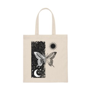 May include: A beige tote bag featuring a black and white butterfly design. The butterfly is bisected, with one half black and the other white. The design also incorporates a sun and moon.