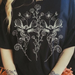 May include: Black t-shirt featuring a detailed white illustration of two winged figures with floral and botanical elements. The design is symmetrical, with the figures facing each other, surrounded by flowers and leaves. The shirt is made of a soft material.