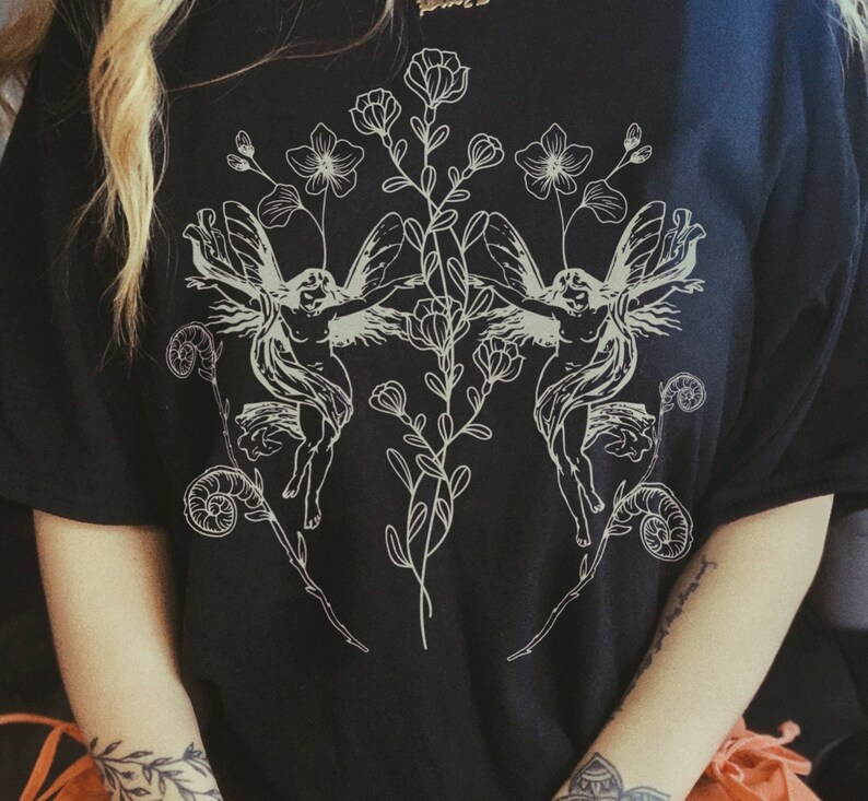 May include: Black t-shirt featuring a detailed white illustration of two winged figures with floral and botanical elements. The design is symmetrical, with the figures facing each other, surrounded by flowers and leaves. The shirt is made of a soft material.