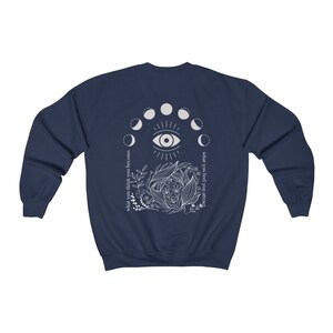 Op de afbeelding: Marineblauw sweatshirt met een wit grafisch ontwerp. Het ontwerp toont een centraal oog, maanfasen, bloemelementen en de teksten "what you think you become" en "what you feel you attract".