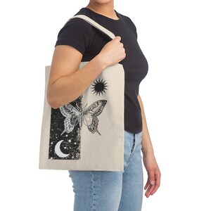 May include: A beige tote bag featuring a black and white butterfly, sun, and moon design. The bag has a black canvas strap.