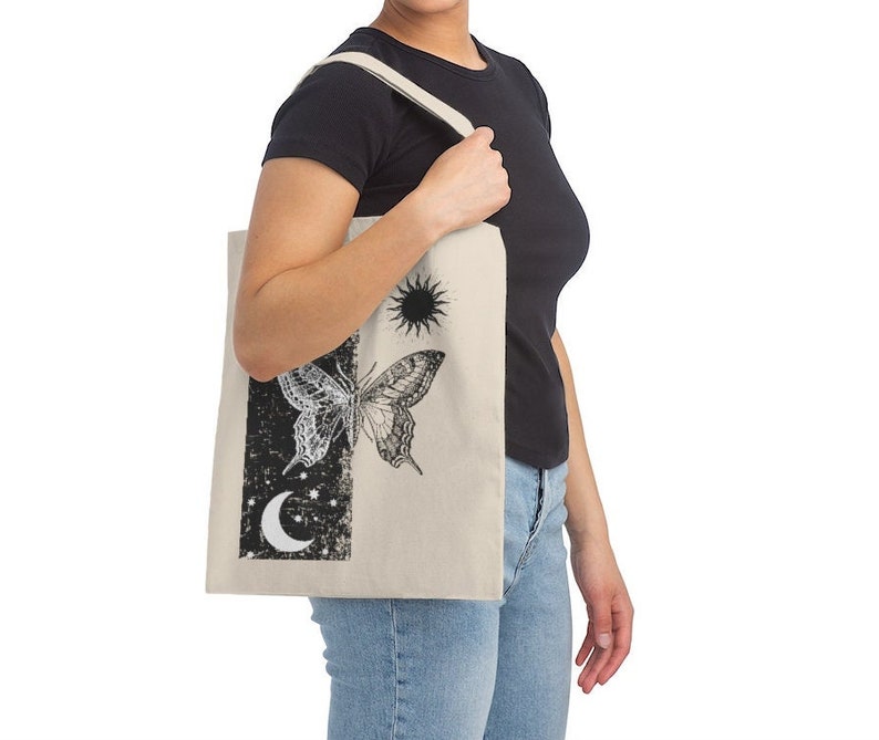 May include: A beige tote bag featuring a black and white butterfly, sun, and moon design. The bag has a black canvas strap.