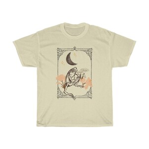 K&ouml;nnte beinhalten: Ein beiges T-Shirt mit einer Vintage-Illustration einer auf einer Blume sitzenden Kr&ouml;te mit einem Halbmond im Hintergrund. Die Illustration ist mit einem dekorativen Rahmen versehen.