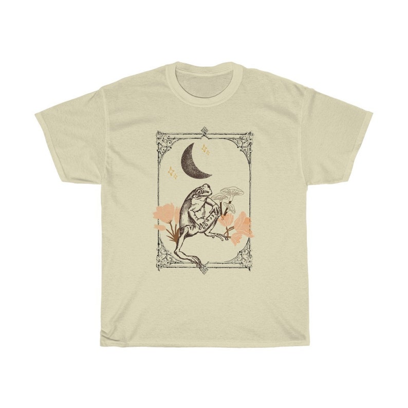 K&ouml;nnte beinhalten: Ein beiges T-Shirt mit einer Vintage-Illustration einer auf einer Blume sitzenden Kr&ouml;te mit einem Halbmond im Hintergrund. Die Illustration ist mit einem dekorativen Rahmen versehen.