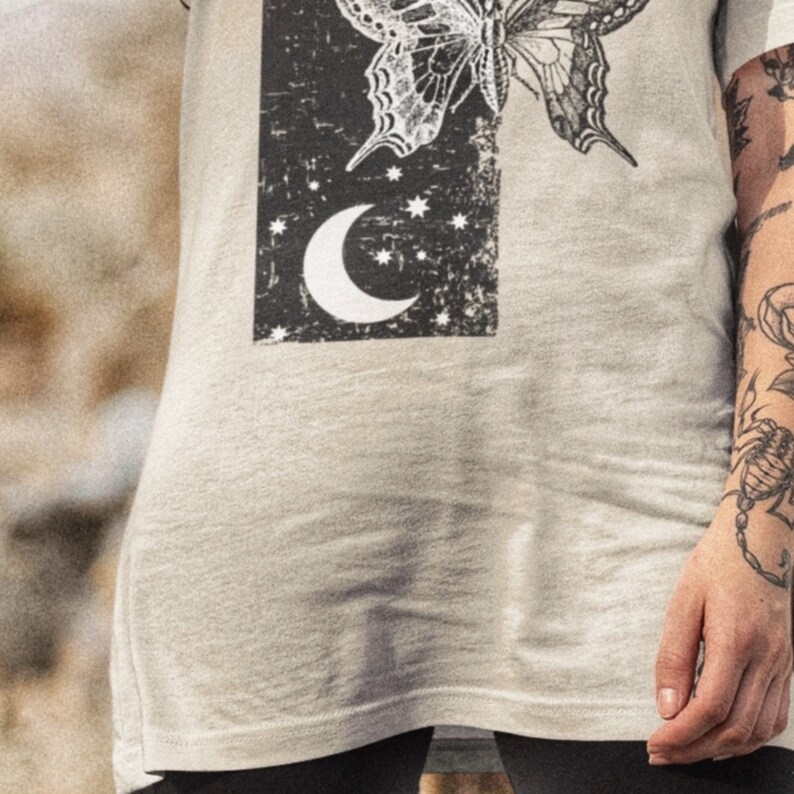 May include: A light gray t-shirt with a black and white graphic print of a butterfly and a crescent moon. The graphic has a vintage, distressed look.