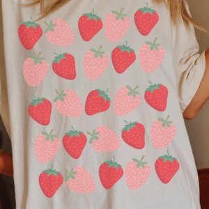 May include: A white t-shirt with a repeating pattern of pink and red strawberries. The strawberries are stylized and have a simple design.