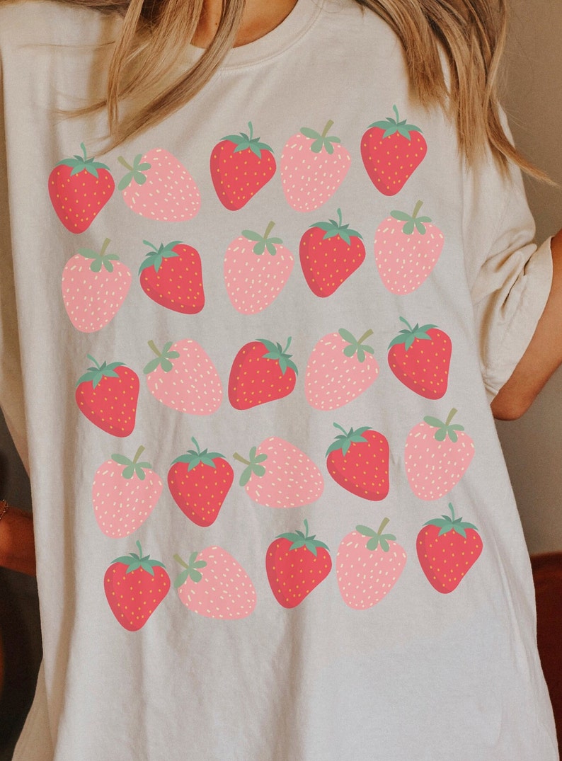 May include: A white t-shirt with a repeating pattern of pink and red strawberries. The strawberries are stylized and have a simple design.