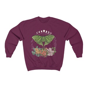 May include: A maroon sweatshirt with a green luna moth design. The moth is surrounded by a circle of white crescent moons and floral accents.