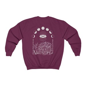 Op de afbeelding: Een bordeauxrode sweatshirt met een wit grafisch ontwerp op de achterkant. Het ontwerp bevat een oog, maanfasen, bloemmotieven en de teksten "what you think you become" en "what you feel you attract".