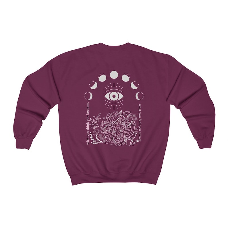 Op de afbeelding: Een bordeauxrode sweatshirt met een wit grafisch ontwerp op de achterkant. Het ontwerp bevat een oog, maanfasen, bloemmotieven en de teksten "what you think you become" en "what you feel you attract".