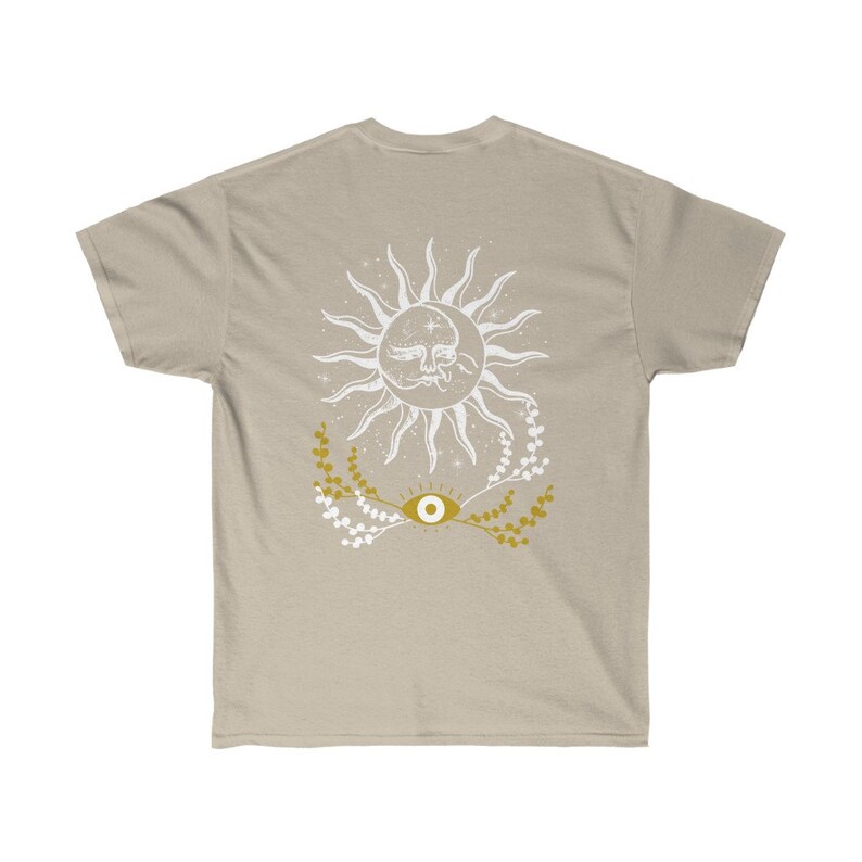 May include: A beige t-shirt with a white sun and eye design. The sun has a face and is surrounded by stars. The eye is gold with white pupils and is surrounded by gold branches.