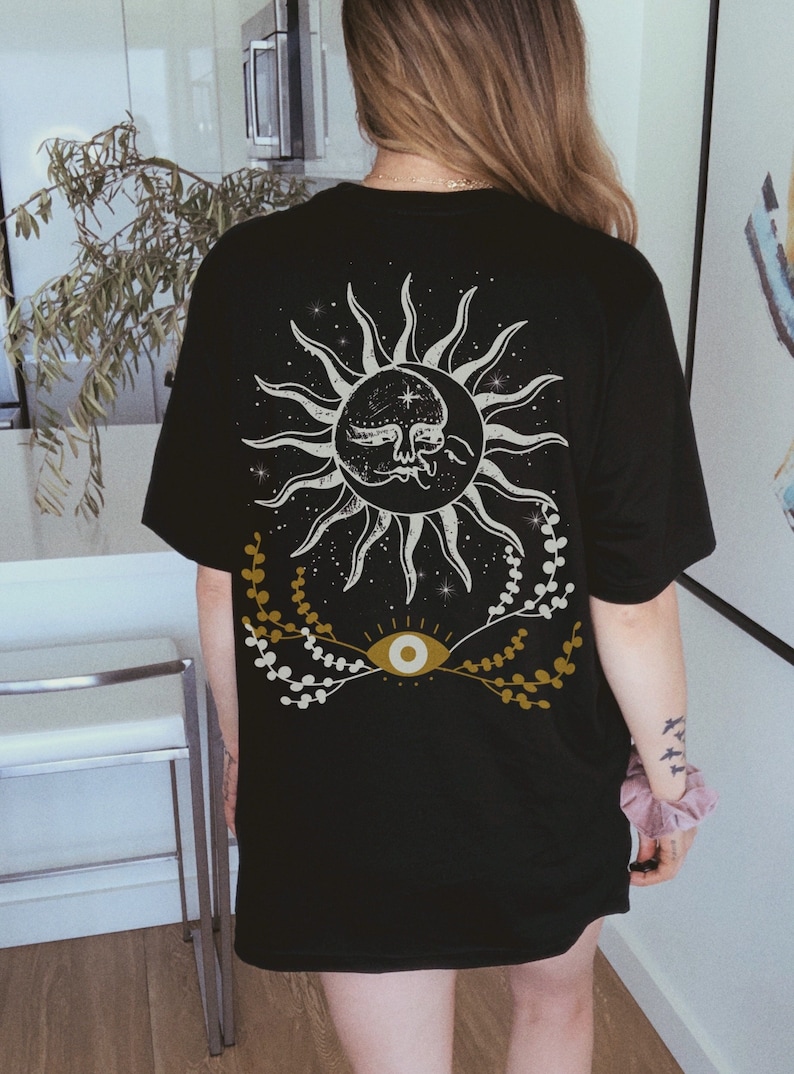 May include: Black t-shirt with a white sun and moon design with a white eye and gold floral accents.
