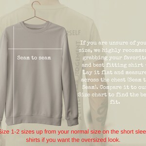 Op de afbeelding: Een lichtgrijs sweatshirt aan een hanger. De afbeelding bevat tekst met meetinstructies, waaronder "Seam to seam". Extra tekst adviseert om een maat groter te nemen voor een oversized look.