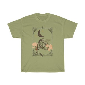 K&ouml;nnte beinhalten: Ein gr&uuml;nes T-Shirt mit einer Vintage-Illustration einer sitzenden Kr&ouml;te unter einem Halbmond mit Sternen und Blumen. Die Illustration ist mit einem dekorativen Rahmen versehen.