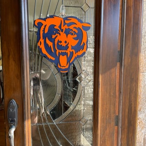 Pu&ograve; includere: Un logo in legno a strati dei Chicago Bears in arancione e blu navy &egrave; appeso a una porta di vetro. La testa dell'orso &egrave; centrata, con la bocca aperta e dettagli. La porta ha un pannello di vetro decorativo e una cornice in legno scuro.