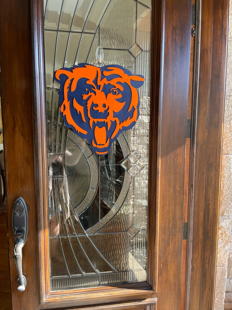Pu&ograve; includere: Un logo in legno a strati dei Chicago Bears in arancione e blu navy &egrave; appeso a una porta di vetro. La testa dell'orso &egrave; centrata, con la bocca aperta e dettagli. La porta ha un pannello di vetro decorativo e una cornice in legno scuro.