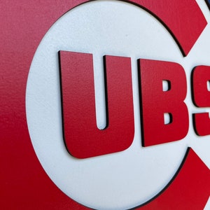 Pu&ograve; includere: Un'insegna in legno con il logo dei Chicago Cubs. L'insegna ha uno sfondo rosso con un cerchio bianco e le lettere "UBS" in rosso. L'insegna &egrave; rifinita in nero e blu.