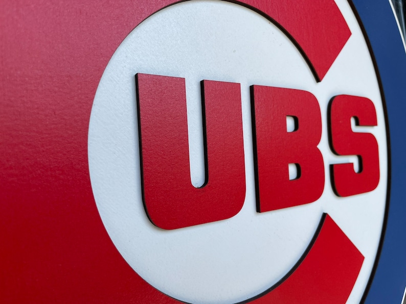 Pu&ograve; includere: Un'insegna in legno con il logo dei Chicago Cubs. L'insegna ha uno sfondo rosso con un cerchio bianco e le lettere "UBS" in rosso. L'insegna &egrave; rifinita in nero e blu.