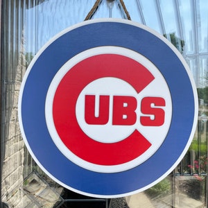 Pu&ograve; includere: Un cartello rotondo in legno con il logo dei Chicago Cubs. Il cartello ha un bordo blu, un cerchio interno bianco e una "C" rossa con la scritta "CUBS" in bianco. &Egrave; appeso a una corda naturale.