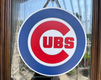 May include: A round wooden sign featuring the Chicago Cubs logo. The sign has a blue outer ring, a white inner ring, and a red "C" with "CUBS" in white letters. A brown rope is attached for hanging.