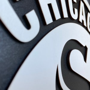 Pu&ograve; includere: Un cartello in bianco e nero con la parola "CHICAGO" in lettere bianche stilizzate. Il cartello ha un design circolare con la lettera "C" in evidenza. Probabilmente si tratta di un oggetto decorativo o di un'opera d'arte murale.