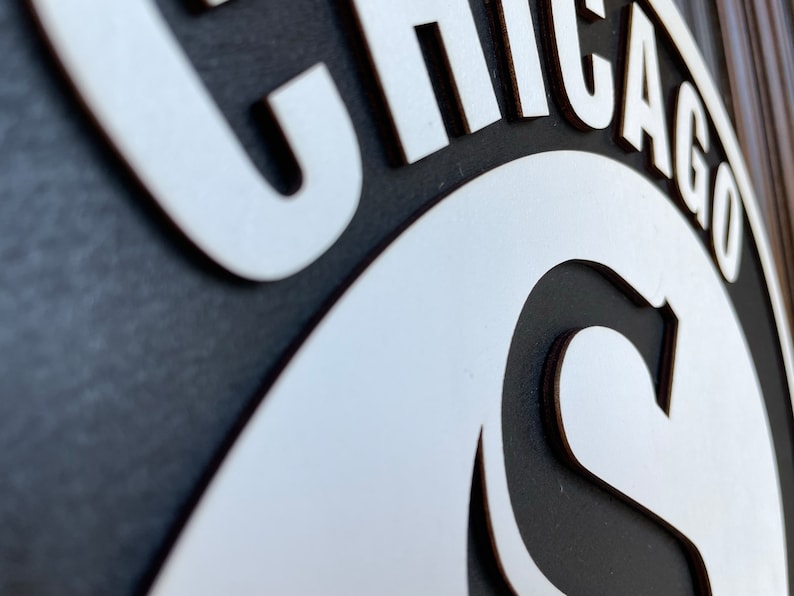 Pu&ograve; includere: Un cartello in bianco e nero con la parola "CHICAGO" in lettere bianche stilizzate. Il cartello ha un design circolare con la lettera "C" in evidenza. Probabilmente si tratta di un oggetto decorativo o di un'opera d'arte murale.