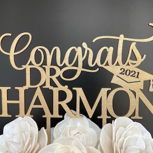 May include: Gold wooden cake topper with the text "Congrats Dr. Harmon 2021" and a graduation cap.