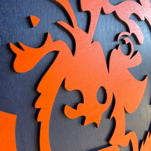 Pu&ograve; includere: Primo piano di un'opera d'arte murale in legno a strati. L'opera presenta un design arancione stilizzato su uno sfondo blu scuro. Il design sembra essere una creatura o un personaggio stravagante.