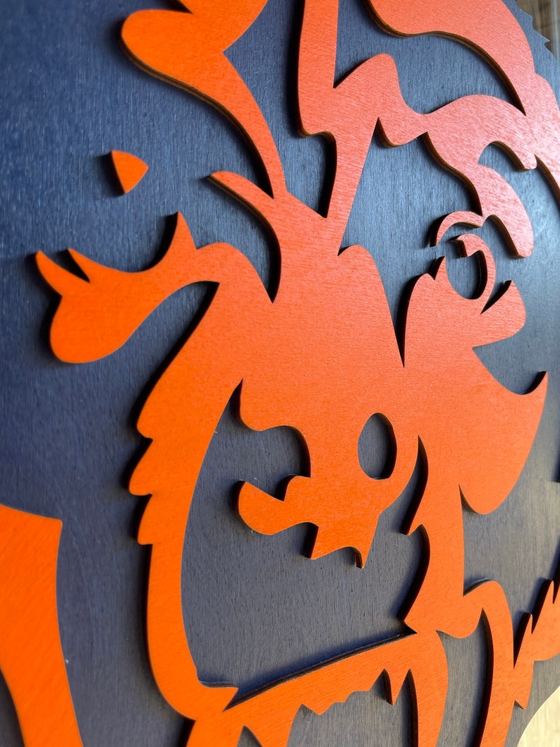 Pu&ograve; includere: Primo piano di un'opera d'arte murale in legno a strati. L'opera presenta un design arancione stilizzato su uno sfondo blu scuro. Il design sembra essere una creatura o un personaggio stravagante.