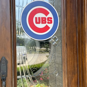 Pu&ograve; includere: Un cartello rotondo con il logo dei Chicago Cubs, con bordo blu, centro rosso e testo bianco "CUBS". Il cartello &egrave; appeso a una porta di vetro con cornice in legno e una corda per appenderlo.
