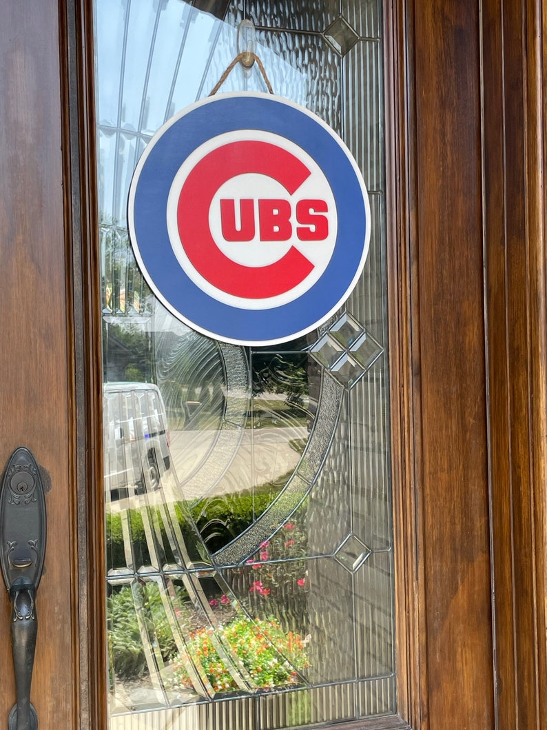 Pu&ograve; includere: Un cartello rotondo con il logo dei Chicago Cubs, con bordo blu, centro rosso e testo bianco "CUBS". Il cartello &egrave; appeso a una porta di vetro con cornice in legno e una corda per appenderlo.