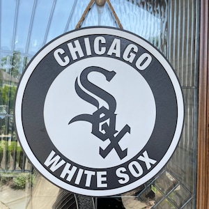 May include: A round, black and white Chicago White Sox team logo sign hanging on a door. The sign features the team's logo in the center, with the words "CHICAGO" and "WHITE SOX" in white lettering around the edge. The sign is made of wood and has a rope for hanging.