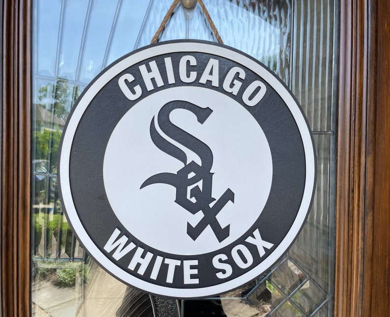 Pu&ograve; includere: Un cartello rotondo dei Chicago White Sox in bianco e nero, appeso a una porta. Il cartello presenta il logo della squadra con le parole "CHICAGO" e "WHITE SOX" in lettere bianche. Realizzato in legno, &egrave; appeso con una corda. Diametro di circa 30 cm.