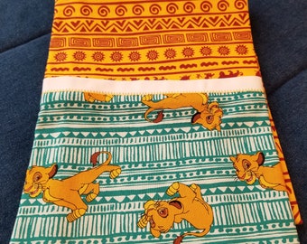 Op de afbeelding: Twee vierkante stoffen met een Lion King-thema. De bovenste vierkant is geel met een rood en wit geometrisch patroon. De onderste vierkant is groen met een wit geometrisch patroon en een herhalend beeld van Simba de leeuwenwelp.