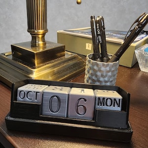 May include: A black and silver perpetual calendar displays "OCT 06 MON". The calendar is on a wooden surface, with a lamp and pen holder in the background. The pen holder contains black pens.