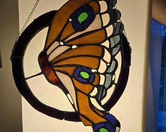 May include: Stained glass butterfly with orange, white, blue, and green details, hanging from a chain. The butterfly is set within a dark brown circular frame. The wings are detailed with various shapes and colors.