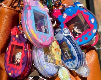 May include: A collection of colorful acrylic keychains featuring cartoon characters and retro handheld game designs. The keychains are in various shapes and colors, including pink, blue, purple, and yellow. They are attached to a silver keyring and are displayed on an orange leather bag.