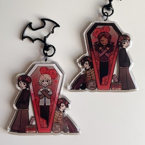 Pode incluir: Dois chaveiros de acrílico com ilustrações de desenhos animados. Um chaveiro tem um topo em forma de morcego. Os chaveiros apresentam personagens em uma moldura em forma de caixão com rosas vermelhas. Os personagens estão em várias poses, com um dentro do caixão.