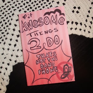 May include: A pink and red card with the words "Awesome Things 2 Do" in a handwritten style. The card also has the phrase "Thats Not The Dang Phone" and a graphic of a phone with a line through it.