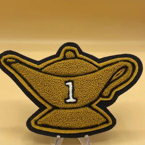 May include: A gold-colored patch in the shape of a genie lamp with a black border. The lamp has a textured surface and a white number 1 in the center. The patch is likely for clothing or accessories.